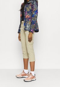 Färgglad mönstrad jacka med långa ärmar, beige croppade byxor och ljusrosa träningsskor med grå accenter och vita strumpor.