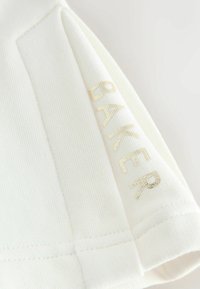 Weißer gerippter Stoffärmel mit goldenem metallischem "BAKER"-Text nahe dem Saum gedruckt, detaillierte Nähte entlang der Kanten sichtbar.