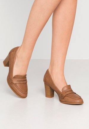 Leren loafers met blokhak in cognackleur, gedragen op blote voeten, staand op een gladde, lichtgekleurde vloer tegen een effen achtergrond.