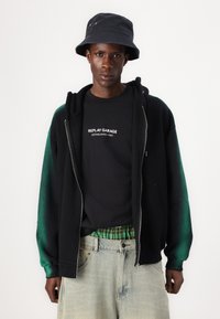 Svart zip-up hettegenser med grønne gradienter på ermene over en svart t-skjorte med hvit tekst. Kombinert med lyseblå denimjeans og svart bøttehatt.