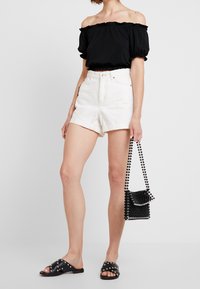 Top negro off-the-shoulder, shorts de denim blancos, sandalias negras con tachuelas y un bolso negro de cuentas. Detalles texturizados y colores contrastantes.