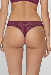 String en dentelle d'un profond bordeaux avec des motifs floraux complexes, texture douce et bords festonnés, présentant un design taille basse.