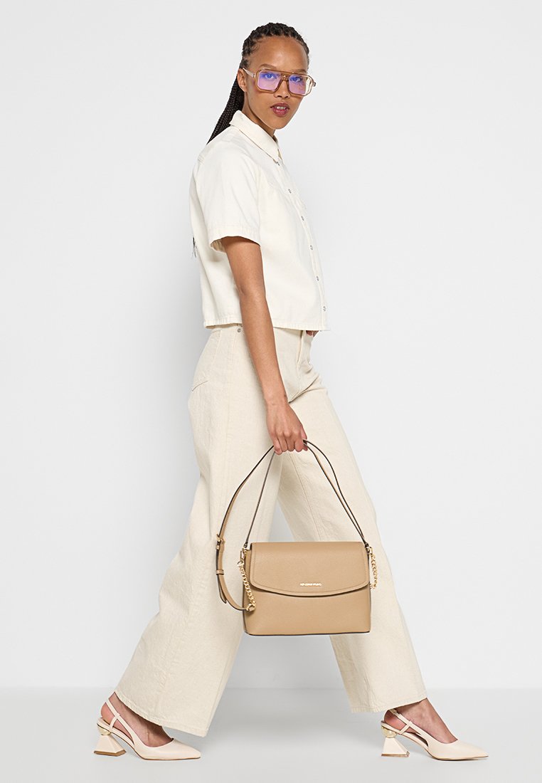 Beige handtas met een gestructureerd ontwerp, een gladde textuur en een gouden kettingdetail. Model draagt een crèmekleurig overhemd met knopen en brede linnen broek.