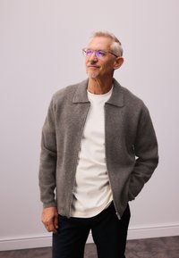 Grijze wollen zip-up jas met een kraag, gedragen over een wit shirt, aangevuld met donkere broek. Het model draagt een bril en heeft kort haar.