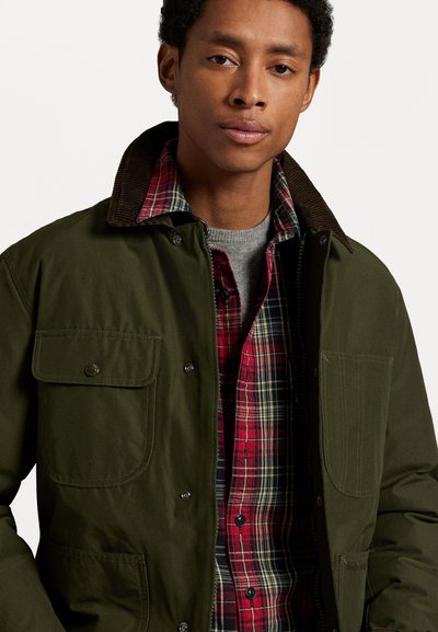 Chaqueta de lona verde oscuro con un cuello de pana, dos bolsillos frontales, superpuesta sobre una camisa de franela a cuadros rojos y un suéter gris de cuello redondo.