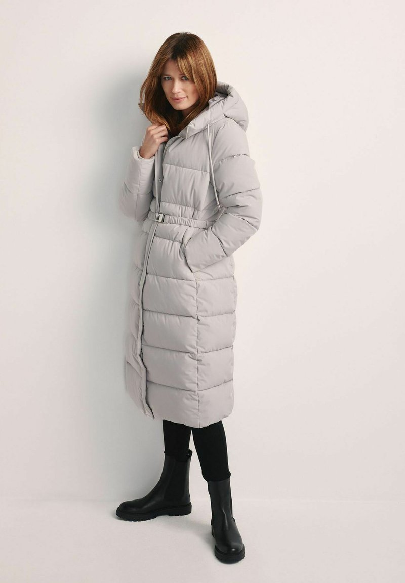 Szara pikowana kurtka puchowa z kapturem, paskiem w talii i przednimi kieszeniami. Charakteryzuje się pikowaną fakturą i długością do kolan, w zestawieniu z czarnymi botkami za kostkę.