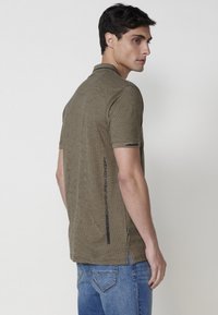 Camisa tipo polo de manga corta verde olivo con un patrón texturizado, aberturas laterales y un acento de marca negra en la espalda. Llevada con jeans azules.