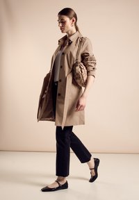 Gabardina beige con botones, jeans negros con dobladillo deshilachado, zapatos planos negros con tiras en el tobillo y un bolso trenzado beige.