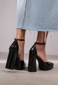 Escarpins en cuir verni noir à talons épais avec des brides de cheville portés avec une jupe en denim bleu clair à ourlet effiloché sur un sol en béton.