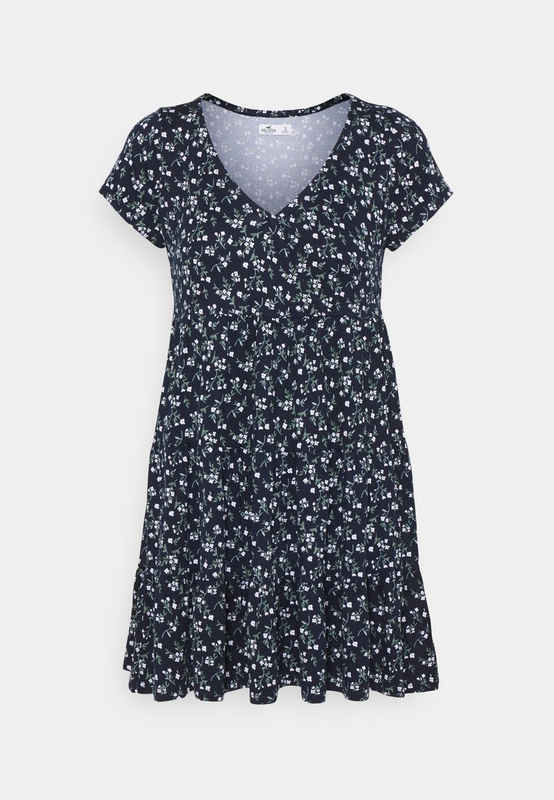 Tummansininen lyhythihainen mekko, jossa on valkoinen kukkakuosi, V-pääntie ja kerroksellinen, liehuva helma.