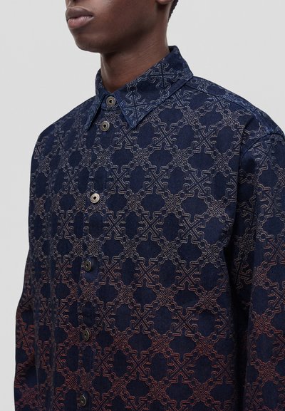 Homme portant une chemise boutonnée avec un motif géométrique de maillons de chaîne bleu et marron et un col pointu, visible du cou jusqu'au milieu du torse.