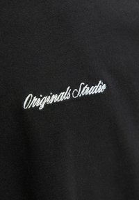 Crna majica s kratkim rukavima od tkanine s bijelim vezom "Originals Studio." Glatka tekstura, ležeran dizajn, izbliza prikaz logotipa.