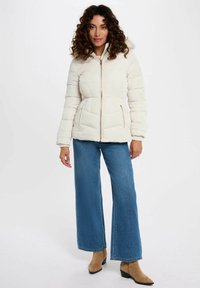Giacca piumino crema con design aderente, cuciture orizzontali e chiusura con zip. Abbinata a jeans a gamba larga blu e stivaletti beige.