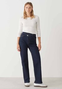 Jeans in denim blu scuro a vita alta con taglio dritto, cinque tasche e cuciture dorate. Abbinati a una camicia bianca a maniche lunghe aderente.