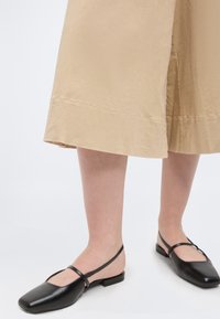 Πόδια που φορούν μπεζ παντελόνια με φαρδιά πόδια και μαύρα επίπεδα παπούτσια slingback με τετράγωνη μύτη και λεπτό λουράκι.