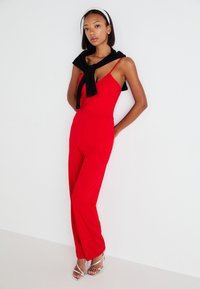 Rød jumpsuit med en tettsittende bodice og vide bukser, parret med en svart genser slengt over skuldrene og sølv høye hæler.