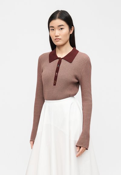 Γυναίκα φοράει ένα μωβ ριμπ πλεκτό μπλουζάκι με μακριά μανίκια, με σκούρο κόκκινο γιακά και κουμπιά, συνδυασμένο με μια λευκή φούστα ψηλόμεση, μπροστά σε απλό φόντο.