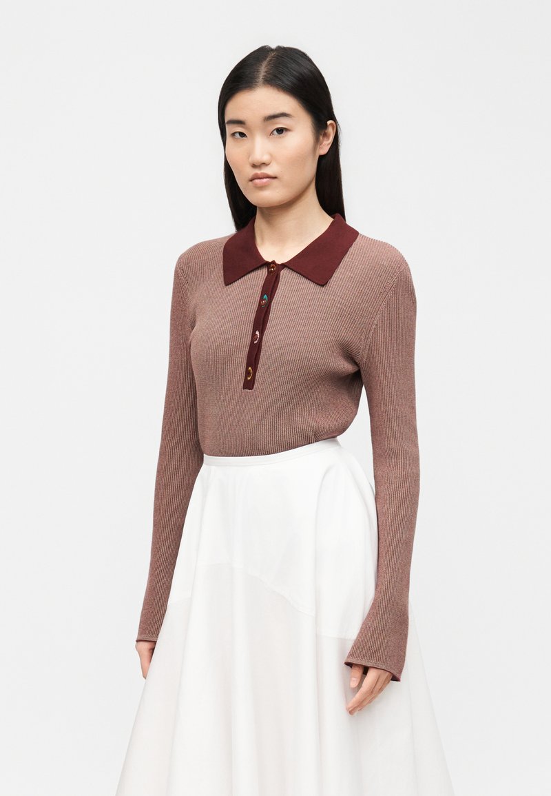 Donna che indossa una maglia a maniche lunghe a coste color malva con colletto e bottoni rosso scuro, abbinata a una gonna bianca a vita alta su uno sfondo neutro.