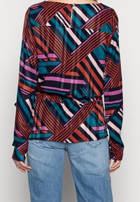 Blouse à manches longues multicolore avec un motif géométrique, présentant des teintes de rouge, bleu et rose, avec une taille cintrée et une fermeture à boutons à l'arrière.