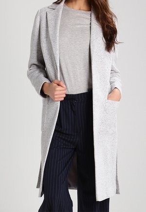 Abrigo largo gris claro en una tela texturizada con solapas de muesca, bolsillos parche y un corte relajado, combinado con una blusa gris y pantalones a rayas.