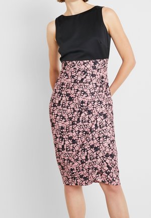 Femme portant un haut noir sans manches avec une jupe crayon taille haute rose à fleurs noires, debout devant un fond uni.