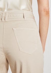 Beige broek met een gladde textuur, voorzien van een achterzak en riemlussen in de tailleband. Eenvoudige stiknaaddetails op de zak.