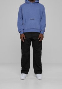 Felpa blu in cotone con tasca frontale e logo "KANI". Abbinata a pantaloni neri e sneaker bianche. Design semplice e casual.