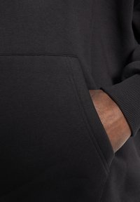 Black sweatshirt with a textured fabric, featuring a front pocket and ribbed cuffs. Close-up shows the hand resting in the pocket.