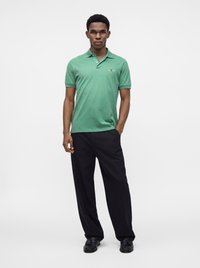 Jeune homme debout portant un polo vert à manches courtes, un pantalon noir à jambes larges et des chaussures noires, sur un fond blanc uni.