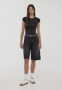 Body aderente nero, pantaloni shorts in denim neri a vita alta e sneakers bianche con suola grigia. I pantaloni shorts hanno una cintura piatta con accenti circolari.