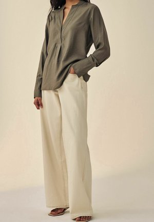Woman wearing olive green long-sleeve blouse with V-neck and cream wide-leg pants, standing with one hand in pocket and sandals visible.