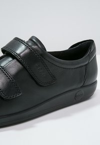 Chaussure à enfiler en cuir noir avec brides ajustables à velcro et semelle en caoutchouc, marquée de logos "ecco" embossés sur la bride et la semelle.
