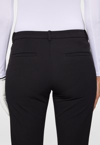 Person som bär åtsittande svarta byxor med bälteshällor och fickor bak, tillsammans med en vit långärmad topp och en vit handske på en hand.