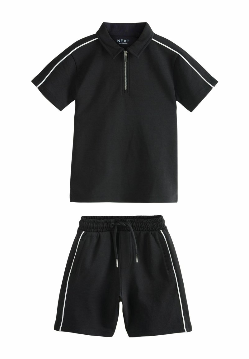 Fekete póló, cipzáros gallérral és fehér oldalsávokkal, párosítva fekete rövidnadrággal, amely rugalmas derékkal és oldalsó zsebekkel rendelkezik.