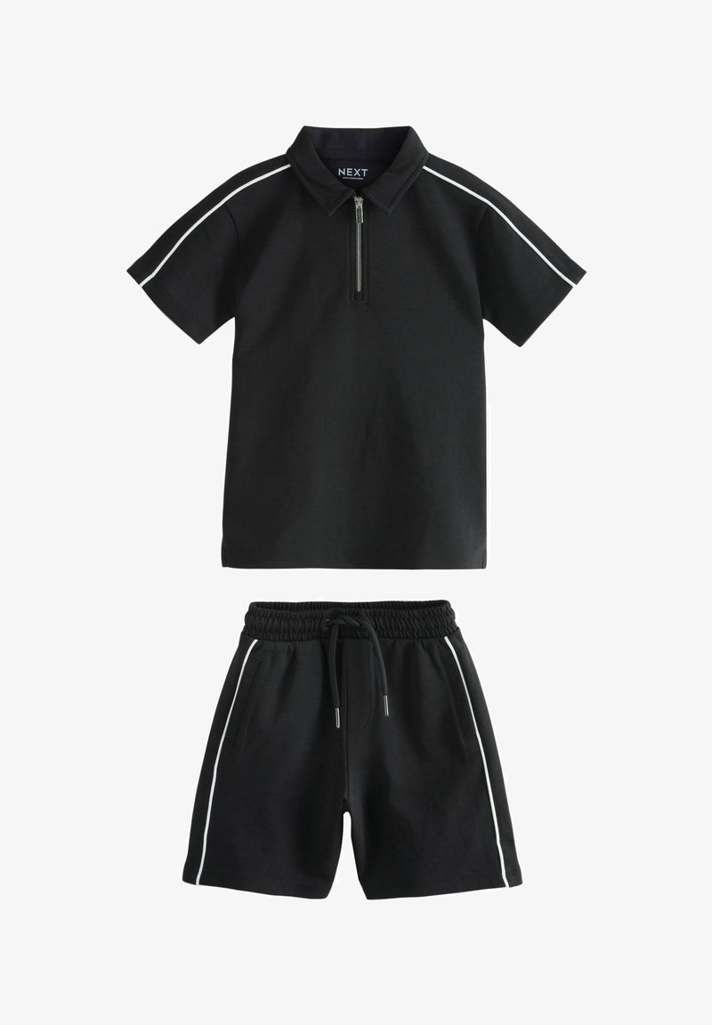 Fekete póló, cipzáros gallérral és fehér oldalsávokkal, párosítva fekete rövidnadrággal, amely rugalmas derékkal és oldalsó zsebekkel rendelkezik.