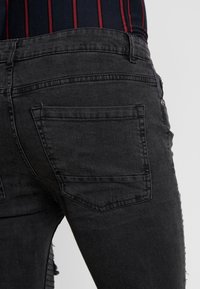 Μαύρες τζιν παντελόνες με στενή εφαρμογή, που διαθέτουν ξεφτισμένα άκρα, σχέδιο πίσω τσέπης και απαλή υφή. Δεν υπάρχουν ορατά λογότυπα ή διακοσμητικά στοιχεία.