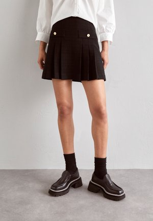 Persona que lleva una falda plisada negra con botones dorados, calcetines negros, zapatos negros gruesos con plataforma y una camisa blanca de manga larga.
