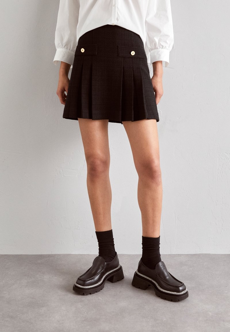 Personne portant une jupe plissée noire avec des boutons dorés, des chaussettes noires, des chaussures compensées noires épaisses et une chemise blanche à manches longues.