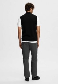 Svart ärmlös stickad väst över en vit t-shirt, tillsammans med gråbyxor och svarta skor, sedd bakifrån.