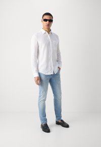 Mann iført svarte solbriller, hvit skjorte med knapper, lyseblå rivebukser og svarte loafers som står mot en enkel hvit bakgrunn.