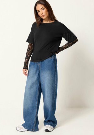Mujer con top negro de manga corta con mangas largas de encaje negro, vaqueros azules de pierna ancha y zapatillas blancas, de pie frente a un fondo claro y liso.