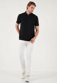 Fekete póló zipzárral a gallérnál, fehér slim-fit farmerral és fehér sneaker cipővel párosítva. A pólónak rövid ujja van és sima textúrája van.