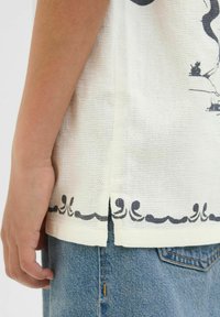 Vit texturerad skjorta med svart vågmönster vid fållen, bärs med blå denimjeans. Skjortan har en sidosprunddetalj.