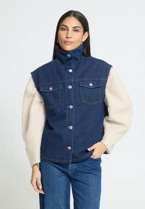 Femme portant un gilet en denim foncé à col montant sur un haut beige clair texturé à manches longues et un jean bleu, debout avec une main dans la poche.