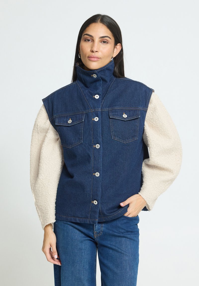 Femme portant un gilet en denim foncé à col montant sur un haut beige clair texturé à manches longues et un jean bleu, debout avec une main dans la poche.