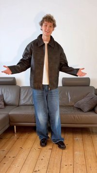 Brązowa skórzana kurtka z szerokim kołnierzem, noszona na białej koszuli. Do kompletu niebieskie luźne dżinsy i czarne buty.