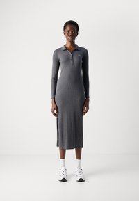 Sivá dlhá polokošeľa s rebrovanou textúrou, postranným rozparkom a vyšitým logom, skombinovaná s bielymi teniskami a ponožkami.