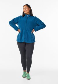 Kvinna stående med händerna på höfterna iförd en blå zip-up jacka, svarta leggings och gröna sneakers framför en vit bakgrund.