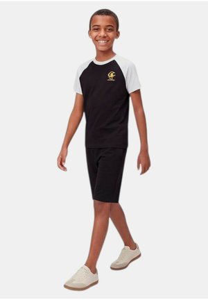 Ragazzo sorridente che cammina, indossa pantaloncini neri, una maglietta con maniche raglan bianco e nero con logo giallo e sneakers bianche.