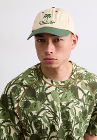 Bežna kapa z zelenim krožnikom, ki vsebuje izvezeno palmovo drevo in besedilo. Zeleno-bež pulover s vzorcem in teksturirano tkanino, krožni ovratnik.